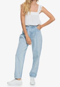 Mujer vestida con una blusa blanca de tirantes con volantes, unos jeans anchos de tiro alto en azul claro y unas zapatillas de plataforma blancas, de pie con una mano en el bolsillo.