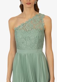Robe vert menthe avec un corsage en dentelle à une épaule à motifs floraux, jupe plissée et ceinture. Détail en dentelle texturée à l'épaule.