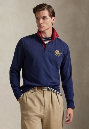 Marineblå langermet polo med rød krage og gult løveemblem. Stripete indre lag. Beige bukser. Mykt materiale, avslappet design.