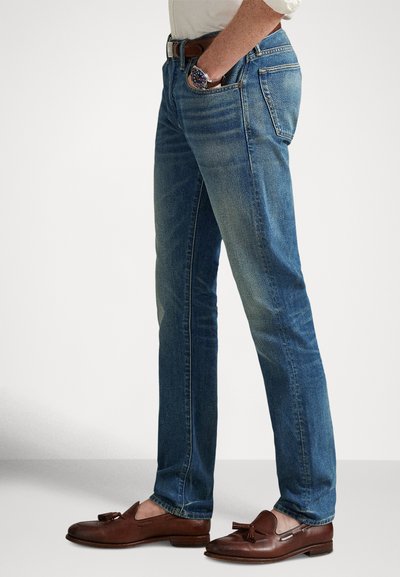 Vīrietis valkā izbalinātas zilas džinsas, baltu kreklu, brūnu ādas jostu, atbilstošas brūnas kurpju ar pušķīšiem, un pulksteni, stāv ar vienu roku kabatā.