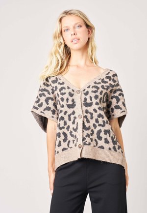 Femme blonde portant un cardigan beige boutonné avec un imprimé léopard noir et un pantalon noir, debout devant un fond clair uni.