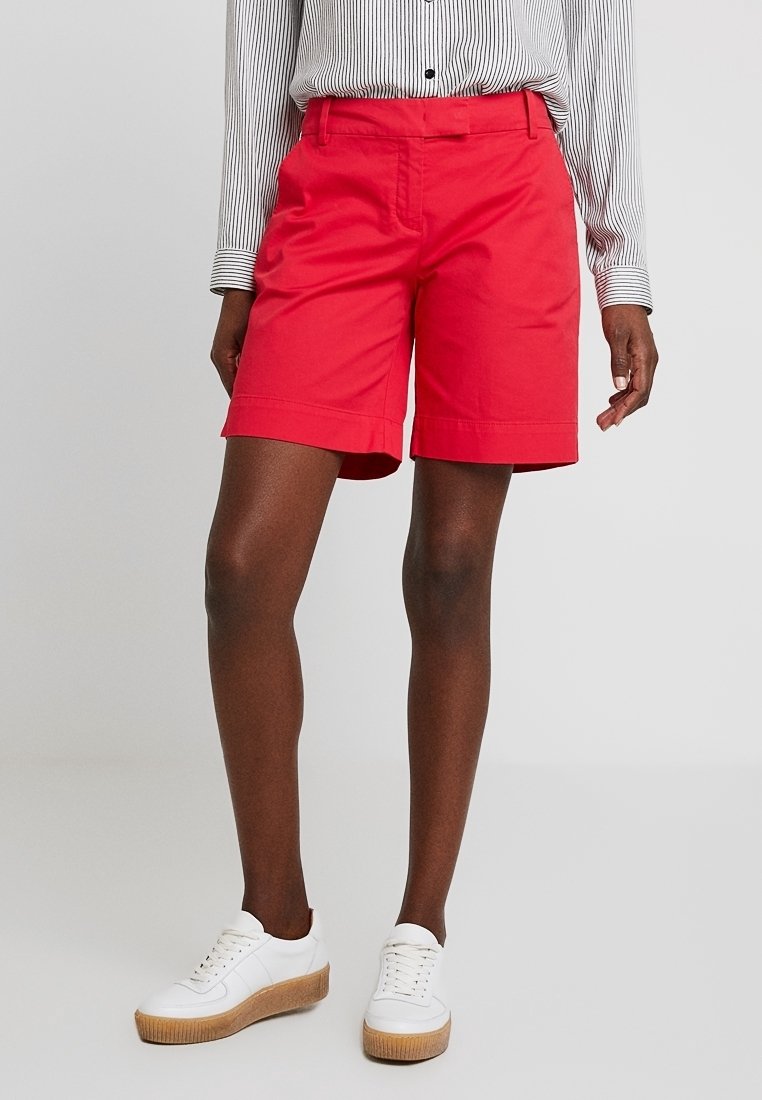 Shorts rouges en coton à l'avant plat, poches latérales et ourlets retroussés, assortis à des baskets blanches avec une semelle épaisse et texturée.