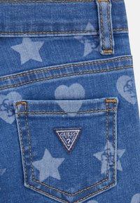 Jeans in denim blu con un motivo a stelle e cuori, dotati di una piccola tasca posteriore e di un'etichetta triangolare con il logo Guess. Sono visibili dettagli cuciti.