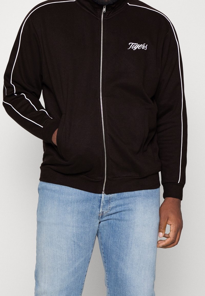 Veste zippée noire avec des rayures blanches le long des épaules, des poches avant et un petit logo "Tigers" sur la poitrine. Tissu en mélange de coton.