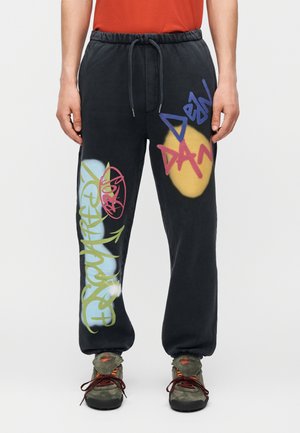 Μαύρη φόρμα με πολύχρωμο κείμενο και σχήματα σε στιλ γκράφιτι, φορεμένη με πράσινα αθλητικά παπούτσια και κόκκινο πουκάμισο, τα χέρια ακουμπούν στα πλευρά.