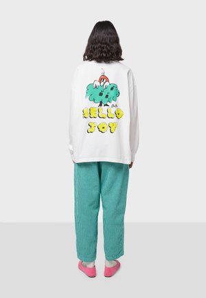 White oversized sweatshirt with colorful print on the back featuring "HELLO JOY" in yellow and a cartoon character. Paired with green striped pants.