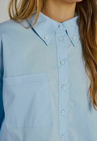 Camicia azzurra chiara con colletto a punta, dotata di taschino sul petto e bottoni bianchi lungo la patta anteriore. Texture di cotone liscia.