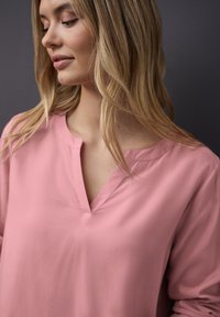Rosa Bluse mit langen Ärmeln, die ein subtiles V-Ausschnitt-Design und eine glatte Textur aufweist. Der Stoff wirkt leicht und fällt sanft.