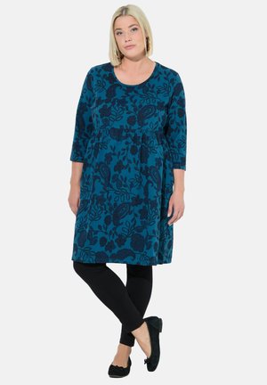 Robe tunique teal avec un motif floral paisley, longueur genou, manches trois-quarts, associée à un legging noir et des chaussures plates noires.