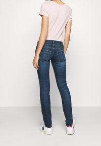 Nainen, jolla on päällään vaaleanpunainen lyhythihainen paita, tummansiniset tiukat farkut ja valkoiset tennarit, kääntää selkänsä yksinkertaista valkoista taustaa kohti.