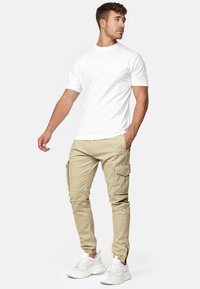 Pantalons cargo beiges avec plusieurs poches latérales, bas ajustés, et un t-shirt à col rond blanc, complété par des chaussures de sport blanches.