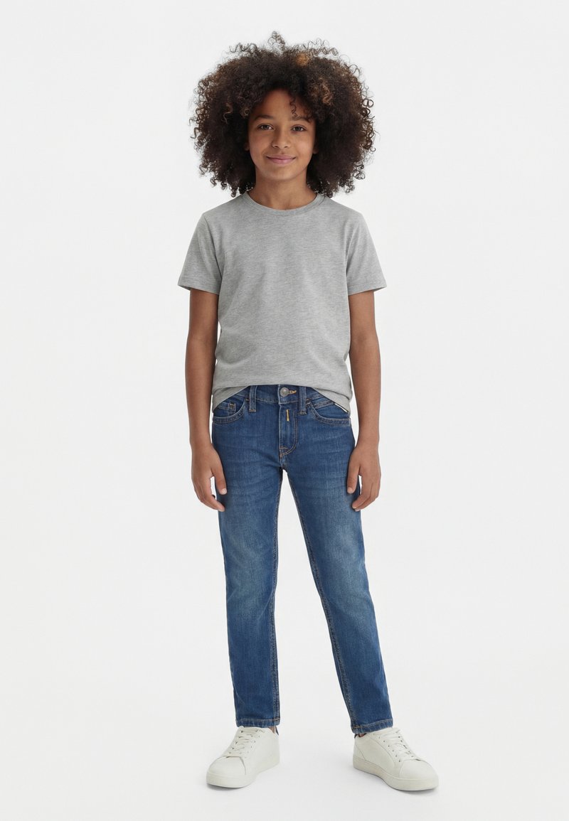 Child with curly hair standing, wearing a grey t-shirt, blue jeans, and white sneakers against a plain white background.