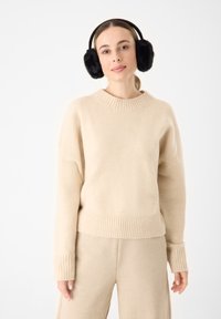 Beige gebreide trui met een geribde onderrand en mouwen, met een ronde halslijn. Gecombineerd met bijpassende beige broek. Zwarte bont oorwarmers.