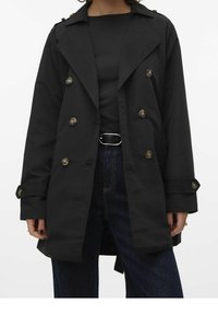 Gabardina negra con botones de doble fila, solapas anchas y bolsillos laterales. Hecha de tela suave. Usada sobre una camiseta negra y jeans oscuros.