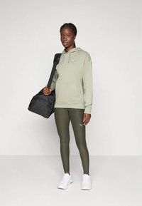 Camisola verde com capuz e leggings oliva em material elástico, combinadas com ténis brancos e uma mala de viagem preta. Design simples com detalhes do logotipo.