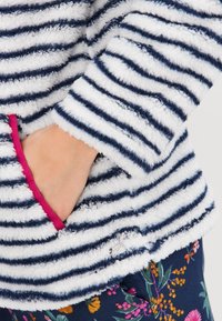 Ręka w kieszeni miękkiej, puszystej kurtki w biało-granatowe paski z różowym wykończeniem, w połączeniu z granatowymi spodniami w kwiatowy wzór.