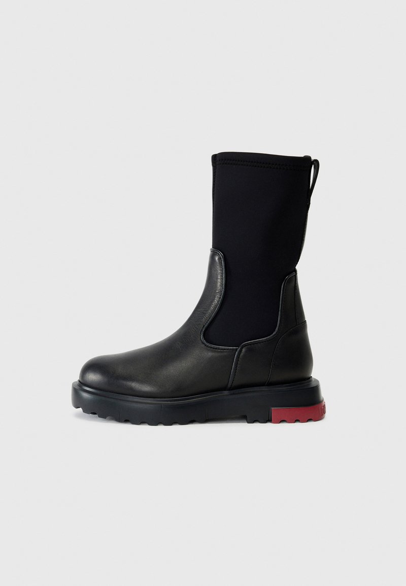 Stivale alla caviglia nero con corpo in pelle liscia, tomaia in tessuto elastico nero e suola in gomma texture con un accento rosso sul tacco.