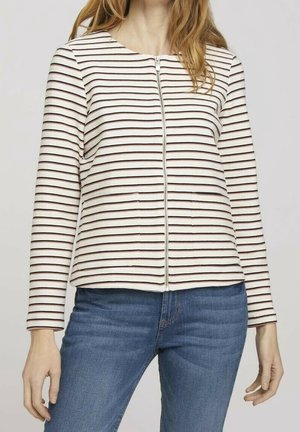 Kvinde iført en hvid og sort stribet zip-up top med lange ærmer og blå jeans mod en ensfarvet baggrund.