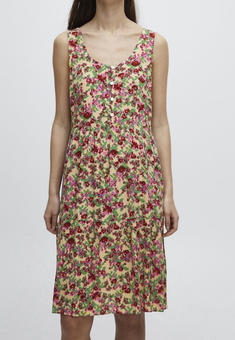 Robe beige sans manches avec motif floral rose et rouge, encolure dégagée, fermeture boutonnée à l'avant et taille froncée portée par une personne.