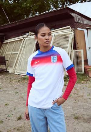 Maillot de football blanc avec des accents rouges et bleus, col rond, et blason de l'Angleterre. Porté par-dessus une chemise à manches longues rouge avec des poignets bleus.