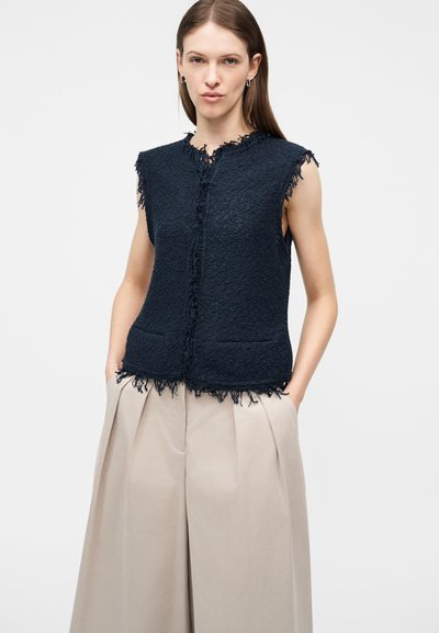 Žena nosi čiernu textúrovanú vesta bez rukávov s ošúchanými okrajmi cez béžové široké skladané nohavice, ruky v vreckách, neutrálne pozadie.