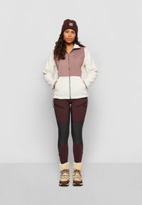 Veste en polaire avec fermeture éclair, présentant un haut rose et un bas crème, assortie à un legging bordeaux et des bottes marron doublées de fourrure.