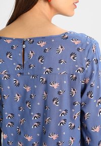 Blusa blu con stampa a mano nei toni di rosa e nero, caratterizzata da una scollatura a chiave sul retro con chiusura a bottone e un tessuto morbido e leggero.