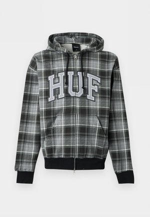 Junger Mann trägt einen schwarz-grau karierten Reißverschluss-Hoodie mit den Buchstaben „HUF“, steht vor einem einfarbigen hellen Hintergrund.