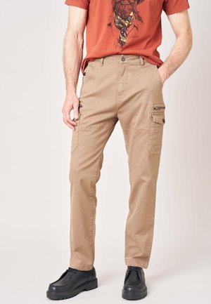 Man draagt beige cargobroek met zijzakken en zwarte schoenen, gecombineerd met een t-shirt in roestkleur met een grafische print.