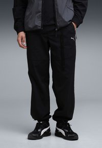 Zwarte atletische joggers met een losse pasvorm, voorzien van een PUMA-logo, ritssluitingen op de zakken en een gestructureerde stof. Gecombineerd met zwarte sneakers.