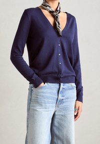 Námornícký cardigan s výstřihem do V, zapínáním na knoflíky a žebrovanými manžetami. V kombinaci se světle modrými džíny. Šátek s černobílým vzorem uvázaný kolem krku.