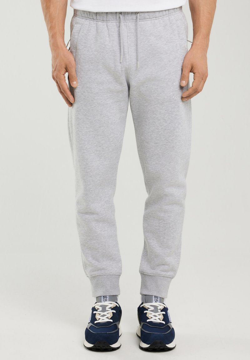 Muž v světle šedých teplákových kalhotách s elastickými manžetami, stahovací šňůrkou v pase a tmavě modrými teniskami s bílými tkaničkami.