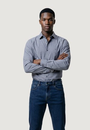 Joven con los brazos cruzados, vistiendo una camisa gris claro abotonada y vaqueros azul oscuro, de pie frente a un fondo claro y liso.