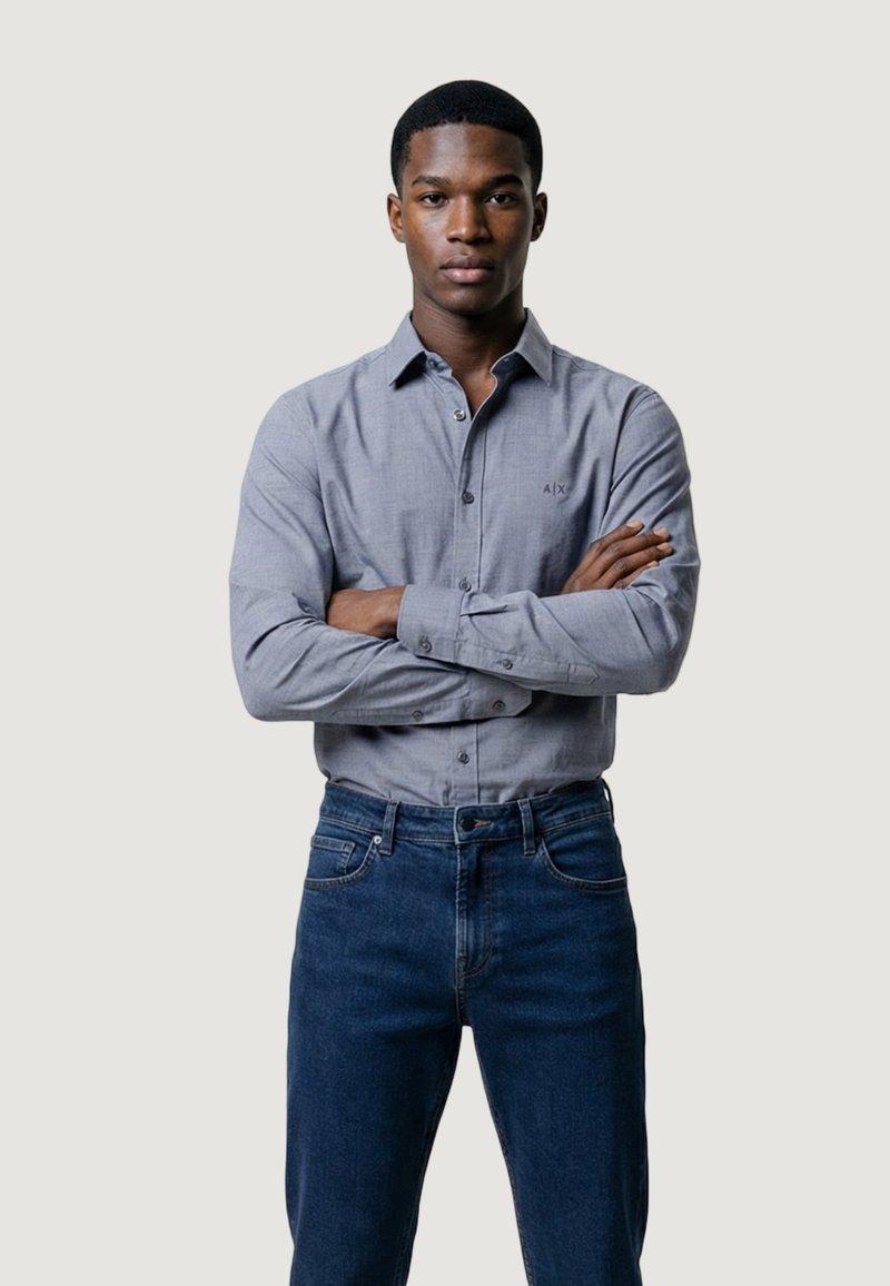 Joven con los brazos cruzados, vistiendo una camisa gris claro abotonada y vaqueros azul oscuro, de pie frente a un fondo claro y liso.