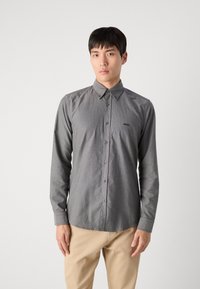 Camisa gris de botones con cuello, mangas largas y una textura sutil. Presenta un pequeño logo negro en el lado izquierdo del pecho.