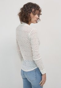 Camicia bianca in pizzo con colletto e maniche lunghe, caratterizzata da un motivo trasparente e testurizzato. Polsini con bottoni e orlo leggermente curvato sul retro.