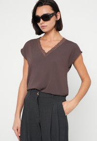 Top marrón de manga corta con escote en V con ribete de encaje, combinado con pantalones plisados gris oscuro. La modelo lleva gafas de sol negras.