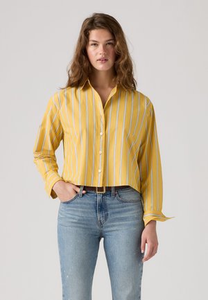 Junge Frau mit welligem Haar, die ein gelbes langärmeliges Shirt mit weißen Streifen und blaue Jeans trägt, steht mit einer Hand in der Tasche.