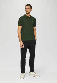 Dunkelgrünes Poloshirt mit kurzen Ärmeln, flachem Kragen und dezentem Logo-Detail, kombiniert mit schwarzen Hosen und weißen Sneakern.