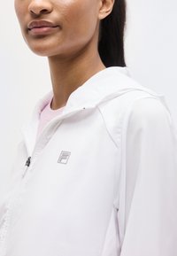 Κοντινό πλάνο ενός ατόμου που φοράει λευκό μπουφάν με φερμουάρ και μικρό λογότυπο "F" στο στήθος, με ροζ ανοιχτό πουκάμισο από μέσα.