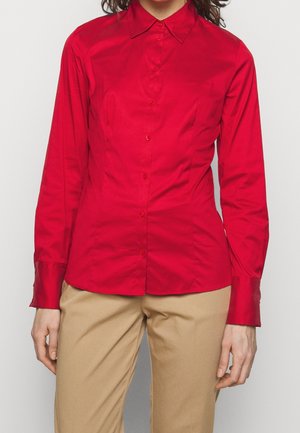 Donna che indossa una camicia rossa aderente con bottoni e maniche lunghe e pantaloni beige, in piedi davanti a uno sfondo chiaro e uniforme.