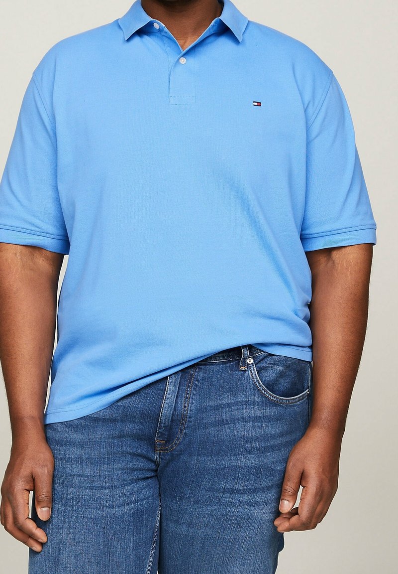 Homme portant un polo bleu clair à manches courtes associé à un jean bleu moyen, debout devant un fond uni.