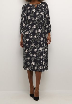 Femme portant une robe midi noire à imprimé floral blanc, manches trois-quarts, ceinture à la taille, et des talons pointus noirs, debout contre un mur uni.