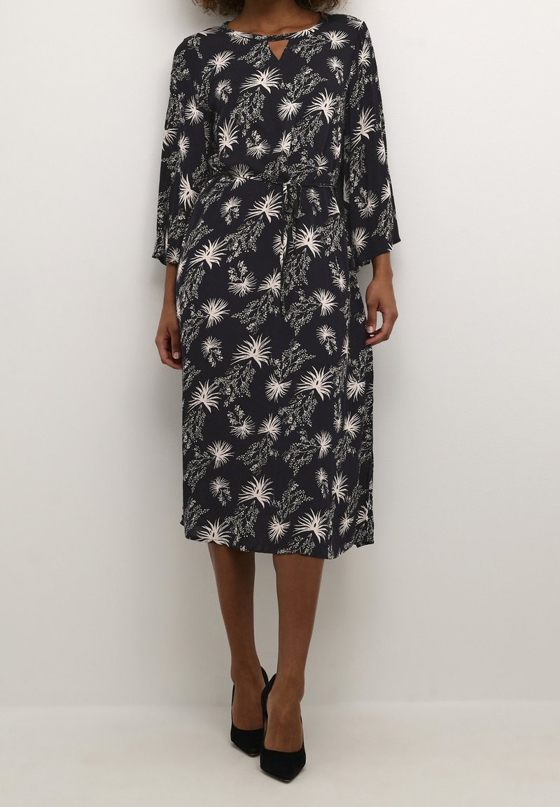 Femme portant une robe midi noire à imprimé floral blanc, manches trois-quarts, ceinture à la taille, et des talons pointus noirs, debout contre un mur uni.