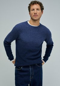 Maglione a coste di colore blu navy con scollatura rotonda, realizzato in morbida lana, presenta maniche lunghe e una vestibilità slim, indossato sopra una camicia bianca.