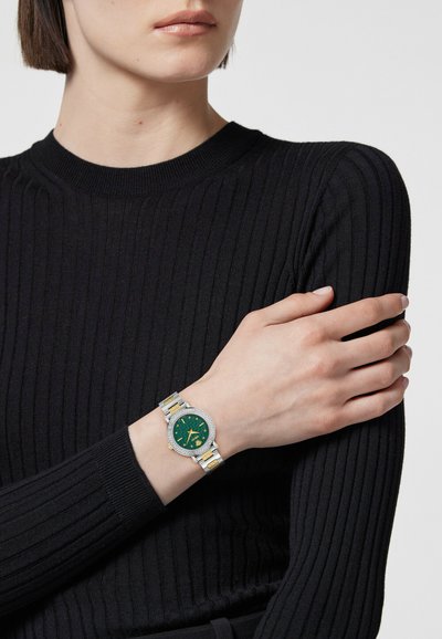 Egy fekete bordázott hosszúujjú felsőt viselő nő, aki ezüst és arany karórát hord zöld számlappal és bonyolult kerettel a bal csuklóján.