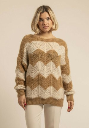Femme aux cheveux blonds courts portant un pull en tricot à motif chevrons marron et crème et un pantalon de couleur claire, debout devant un fond uni.