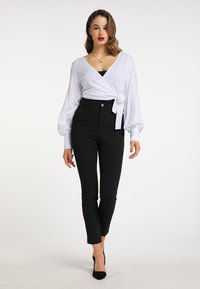 Weiße Wickelbluse mit langen, aufgestellten Ärmeln und einer Schleife in der Taille, kombiniert mit schwarzen hochtaillierten Hosen und schwarzen spitzen Pumps.