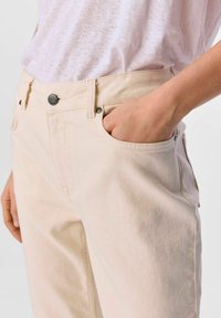 Vale beige puuvillahousut, joissa on napillinen vyötärö, etutaskut ja sileä pinta, kuvattuna käsi taskussa.