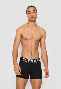 Crne bokserice s bijelim pojasom s natpisom "CALVIN KLEIN", s prilagođenim dizajnom i glatkom teksturom tkanine.